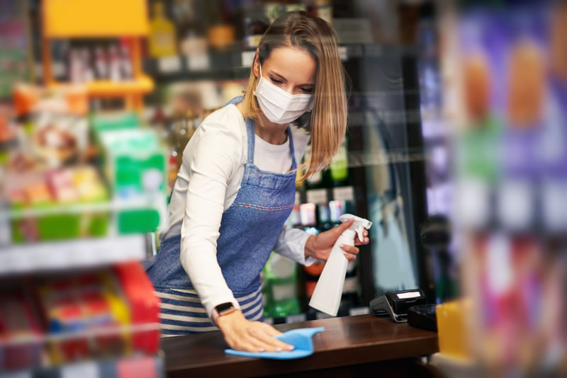 storecleark-cleaning-store A female store clerk in an apron and mask wipes down a counter.